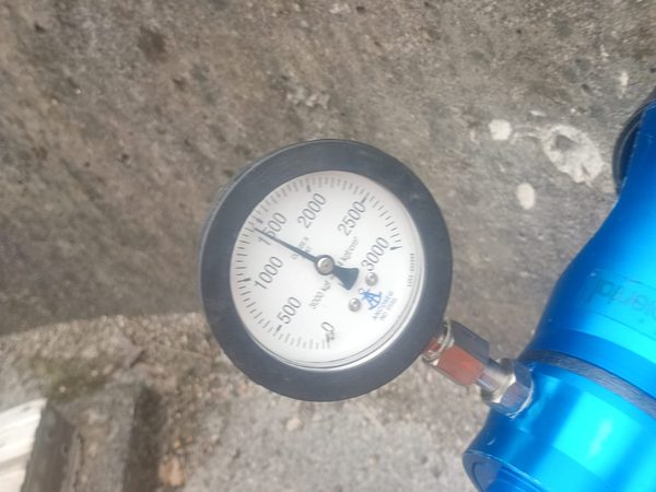 Manômetro analógico com escala graduada em kgf/cm² acoplado por conexão metálica ao cilindro hidráulico azul do equipamento de ensaio